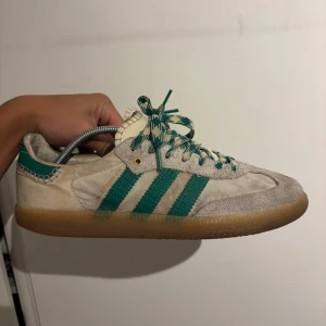 Adidas Samba Wales Bonner sneakers - Adidas Samba Wales Bonner sneakers i beige mocka och textil med gröna detaljer och klassiska tre ränder. Skorna har unika mönstrade snören i grönt och vitt samt gummisula. Wales Bonner-logga i guld på sidan och snygga kontrastsömmar vid häl och plös. Passar 41