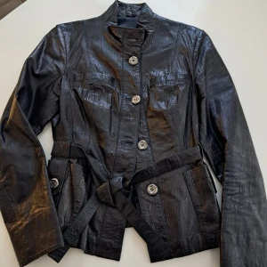 Svart skinnjacka från Esccada exklusiv  - Snygg svart skinnjacka från Esccada Exclusive i storlek S. Jackan har markerad midja med bälte, stora knappar framtill och långa ärmar. Den har en cool, lite glansig finish och stilren design som ger en edgy vibe. Perfekt för dig som gillar statement-plagg och vill sticka ut.