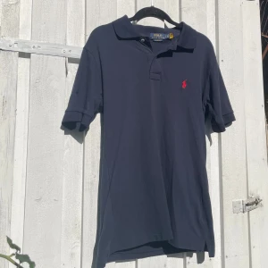 Marinblå pikétröja från Ralph Lauren - Snygg marinblå pikétröja från Polo Ralph Lauren med klassisk krage, två knappar och röd broderad logga på bröstet. Tillverkad i bomull och har korta ärmar. Perfekt för en stilren och avslappnad look.