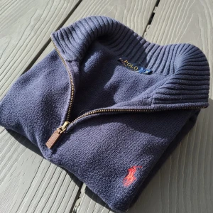 Blå stickad tröja från Polo Ralph Lauren - Säljer en snygg blå stickad tröja från Polo Ralph Lauren med dragkedja och ribbad krage. Den har en liten röd logotyp på bröstet och ribbade muddar vid ärmslut och nederkant. Perfekt inför sommarn!