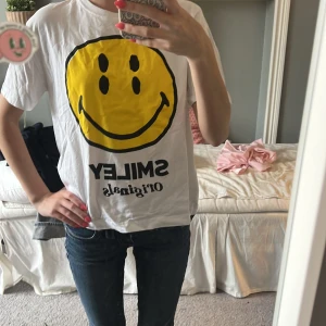 Vit Smiley Originals t-shirt - Vit t-shirt från Zara med stor gul smiley på framsidan och svart text både fram och bak. Klassisk passform med korta ärmar och rund hals. Perfekt statement-plagg för dig som gillar coola tryck och streetstyle.