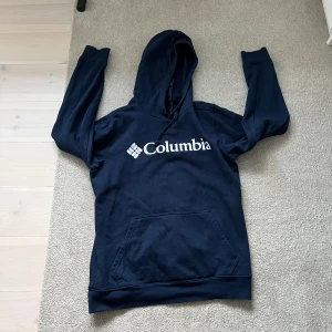 Mörkblå hoodie från Columbia - Snygg mörkblå hoodie från Columbia med stor vit logga på bröstet. Tröjan har huva med snörning och en klassisk känguruficka framtill. Perfekt för kyliga dagar och chill outfits.