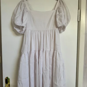 Vit puffärm klänning med knyt i ryggen - Superfin vit klänning med puffärmar och volanger. Den har fyrkantig urringning och snygg knytning i ryggen som ger en öppen rygg-effekt. Perfekt för varma dagar och har en luftig, flowy passform. 