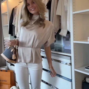 Beige blus med markerad midja - Säljer en stilren beige blus med lätt struktur i tyget och markerad midja. Blusen har korta, vida ärmar och en mjuk, rundad halsringning. Perfekt att styla med jeans eller kjol för en clean look.