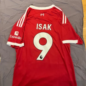 Liverpool  fotbollströja Isak  - Röd Liverpool fotbollströja från Adidas med vita detaljer och tryck. Nummer 9 och namnet Isak på ryggen, Premier League-logga, Standard Chartered och Expedia som sponsorer. Klassisk passform, kortärmad och tillverkad i lätt funktionsmaterial.