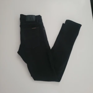 Svarta jeans från Nudie Jeans, 28/30 - Svarta jeans från Nudie Jeans med klassisk femficksdesign och diskret logga på bakfickan. Jeansen har en smal passform och är tillverkade i mjukt bomullstyg med stretch för extra komfort. Perfekta för dig som gillar stilrena och enkla svarta jeans.