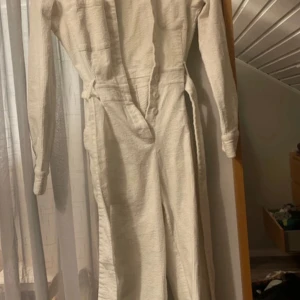 Vit manchester jumpsuit från NA-KD - Säljer en snygg vit jumpsuit från NA-KD i mjuk manchester. Den har långa ärmar, knappar framtill, två bröstfickor och ett bälte i midjan för extra stil. Perfekt för dig som gillar en clean och cool look.