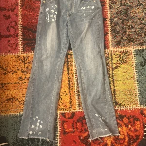 Ljusblå jeans med slitna detaljer - Säljer ett par ljusblå jeans med raka ben och coola slitningar samt färgstänk över hela byxan. Jeansen har råa, fransiga benslut och klassisk femficksdesign. Perfekta för dig som gillar en avslappnad och trendig streetstil. Dessa är i flared stil/ streetwear och köptes för 500 kr säljer nu för 350 pris kan diskuteras 