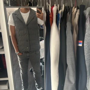 Grå rutig dunväst från Slowear - Stilren grå dunväst från Slowear med diskret rutigt mönster. Västen har hög krage, knäppning med knappar framtill och quiltad design. Yttermaterialet är i ullblandning som ger en exklusiv känsla och passar perfekt till lager-på-lager-stil.