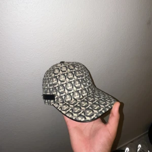 Salvatore Ferragamo mönstrad keps - Snygg keps från Salvatore Ferragamo med grått och beige mönster över hela kepsen. Justerbart spänne bak och svart läderdetalj med logga. Tillverkad i bomull med svart innerfoder. Perfekt accessoar för en trendig look.