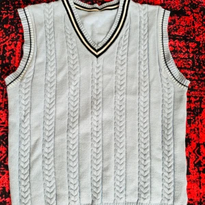 Ljus stickad kabelväst med v-ringning - Kabelstickad väst i ljus färg med v-ringad hals och svarta/vita ränder runt hals och ärmöppningar. Klassisk preppy stil, perfekt att bära över skjorta eller t-shirt. Mjuk och skön stickad känsla.