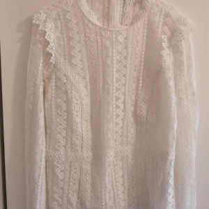 Vit blus med lång ärm - Superfin vit blus i spets med lång ärm och högt rundat halsringning. Blusen har vackra spetsdetaljer, mönster med både zigzag och prickar samt helfodrad insida. Perfekt för dig som gillar romantisk och stilren look. Har endast använts ett fåtal gånger. 