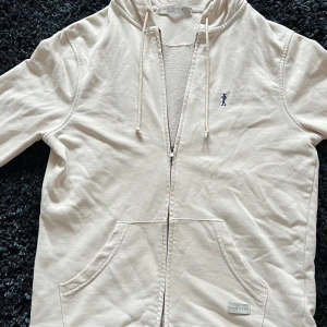 Vit hoodie från Polo Club - Snygg vit hoodie från Polo Club med dragkedja, huva med snörning och två stora fickor framtill. Broderad blå logga på bröstet. Tillverkad i mjuk bomull, perfekt för chill dagar. Klassisk och enkel design som funkar till det mesta. Använt ett par gånger, men inget som påverkat utseendet.