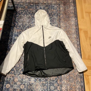 Nike svartvit vindjacka med huva - Nike vindjacka i svart och vitt med snygg kontrastdesign. Jackan har huva, hel dragkedja framtill och två fickor med dragkedja. Lätt och sportig känsla, perfekt för blåsiga dagar. Nike-logga på bröstet och meshfoder på insidan för extra komfort. Inga skador.