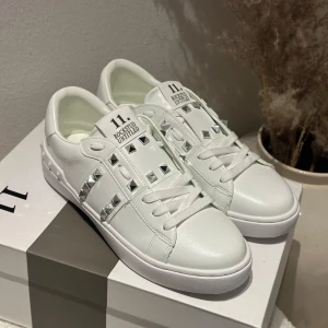 Valentino Rockstud vita sneakers - Snygga vita Valentino Garavani Rockstud sneakers i skinn med ikoniska silverfärgade nitar och platt sula. Klassisk rund tå, snörning och coola detaljer längs sidor och häl. Perfekta för dig som vill sticka ut med en lyxig touch.