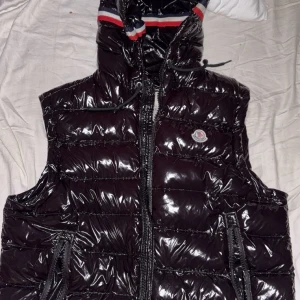 Svart dunväst från Moncler med huva - Snygg svart dunväst från Moncler med glansig finish och quiltad design. Västen har huva med röd, vit och blå rand, dragkedja framtill och två fickor med dragkedja. Klassisk Moncler-logga på bröstet. Perfekt för lager-på-lager under kyliga dagar.