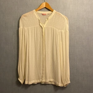 Vit prickig blus från Indiska - Luftig och flowy vit blus från Indiska med diskreta prickar i tyget. Blusen har v-ringning, knappar framtill och långa ärmar med lätt rynk vid axlar och ärmslut. Perfekt för en chill och stilren look.