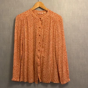 Mönstrad orange skjorta från Indiska - Säljer en snygg orange skjorta från Indiska med vitt blommigt mönster. Skjortan har långa ärmar, rund halsringning och knappar framtill. Perfekt för dig som gillar färg och mönster!