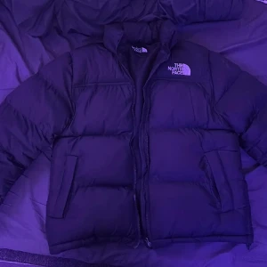 Svart pufferjacka från The North Face - Säljer en svart pufferjacka från The North Face med klassisk logga på bröstet och ryggen. Jackan har hög krage, dragkedja framtill och två sidofickor. Perfekt för kalla dagar och riktigt snygg till streetwear-stilen.