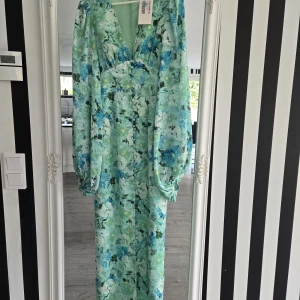 Mintgrön blommig långklänning Adoore 32 - Superfin långklänning från Adoore i mintgrönt med blått och vitt blommönster. Klänningen har v-ringning, långa puffärmar och knappar framtill. Tillverkad i ett lätt och luftigt material, perfekt för sommarens alla tillfällen.