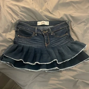 Mörkblå volangkjol i jeans från Hollister - Supersöt mörkblå jeanskjol från Hollister med volang och råa kanter. Klassisk femficksmodell med snygga kontrastsömmar och broderade bakfickor. Kjolen har en tight passform upptill och voluminös volang nertill för extra edge. (Sydde den själv) 