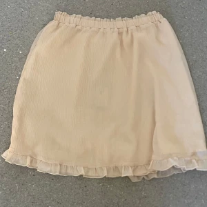 Beige kortkjol med volangkant - Söt beige kortkjol med volangdetalj längst ner och resår i midjan. Kjolen har en lätt och luftig känsla, perfekt för varmare dagar. Enkel att matcha med olika toppar för en trendig look.