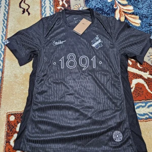 Svart AIK x Nike fotbollströja 1891 - AIK x Nike fotbollströja i svart med diskret mönster, klubbmärke på bröstet och trycket '1891' framtill. Kortärmad modell i lätt och ventilerande material. Detaljer som 'SMOKINGLIRARE' på ryggen. Perfekt för dig som vill representera AIK med stil.