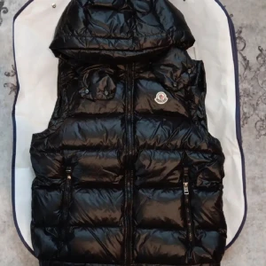 Svart dunväst från Moncler med huva - Snygg svart dunväst från Moncler med glansig finish, huva och två dragkedjefickor framtill. Västen har quiltad design och klassisk Moncler-logga på bröstet. Perfekt för lager-på-lager och streetstyle.