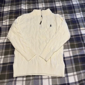 Vit stickad half zip-tröja Polo Ralph Lauren - Vit kabelstickad tröja från Polo Ralph Lauren med half zip och broderad logga på bröstet. Tröjan har ribbade muddar och hög krage, perfekt för lager-på-lager. Klassisk och stilren design som funkar året runt.