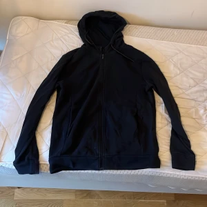 Svart hoodie från Dressmann i bomull - Svart hoodie från Dressmann i storlek M. Tröjan har dragkedja framtill, huva och långa ärmar. Tillverkad i mjuk bomullsblandning med 8% polyester för extra komfort. Perfekt för en chill och avslappnad stil.
