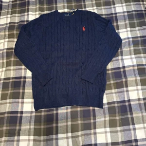 Mörkblå kabelstickad tröja Polo Ralph Lauren - Kabelstickad mörkblå tröja från Polo Ralph Lauren med klassisk röd broderad logga på bröstet. Tröjan har rund halsringning, ribbade muddar och är långärmad. Perfekt för dig som gillar stilren och tidlös design.
