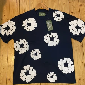 Blå blommig t-shirt från Denim Tears - Mörkblå t-shirt från Denim Tears med stora vita blommiga tryck över hela plagget. Klassisk passform och rund halsringning. Tillverkad i 100% bomull för en skön känsla. Perfekt för dig som vill sticka ut med en unik och trendig look.