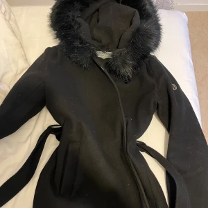 Svart kappa med fuskpäls från Hollies - Säljer en svart kappa från Hollies med stor huva och fluffig fuskpäls runt kanten. Kappan har bälte i midjan, raka fickor och en snygg guldig detalj på ärmen. Perfekt för kalla dagar när du vill vara både varm och stilren. Köpte för 1899kr . Kan diskutera priset om behövs 🤍