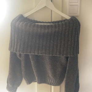 Mörkgrå offshoulder stickad tröja - Säljer en mörkgrå stickad tröja med bred ribbad offshoulder-krage. Tröjan har lång ärm och en loose passform, perfekt för kyliga dagar. Den stickade strukturen ger en mysig känsla och den breda kragen gör den extra trendig.
