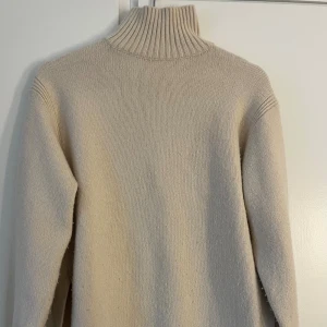 Beige polotröja med slits i sidan - Beige stickad polotröja med ribbad hög krage och långa ärmar. Tröjan har en snygg slits i sidan och breda ribbade muddar vid ärmslut och nederkant. Perfekt för lager-på-lager under kyliga dagar.