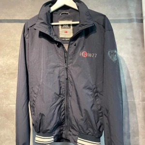 vindjacka Helly Hansen - Säljer en mörkblå vindjacka från Helly Hansen med broderad logga och norsk flagga på bröstet samt emblem på ärmen. Jackan har dragkedja framtill, ribbade muddar med vita ränder och ståkrage. Perfekt för blåsiga dagar och snygg till jeans.