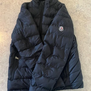 Moncler - Säljer en svart pufferjacka från Moncler med diskret logga på ärmen. Jackan har dragkedja framtill och två sidofickor med dragkedja. Klassisk quiltad design och hög krage som skyddar mot vind. Perfekt för kyliga dagar.                  