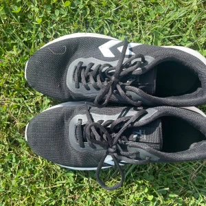 Svarta sneakers från Hummel - Svarta sneakers från Hummel med vit sula och vita detaljer på sidorna. Ovandelen är i mesh-liknande syntetmaterial och skorna har snörning. Perfekta för dig som gillar sportig stil och vill ha bekväma skor till vardags.