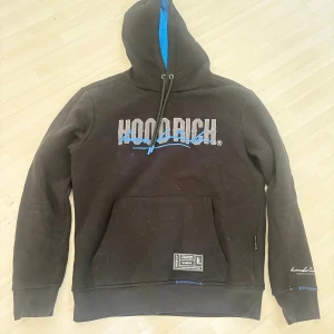 Svart hoodie från Hoodrich med blå detaljer - Svart hoodie från Hoodrich med broderad logga i grått och blått framtill och baktill. Tröjan har huva med blå insida, snörning och en stor magficka. Extra detaljer som etikett nertill och broderad text på ärmen.