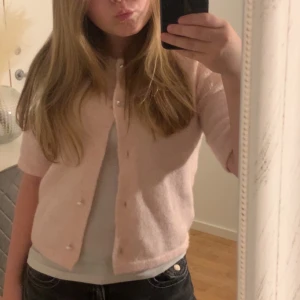 Ljusrosa kofta med pärlknappar - Supersöt ljusrosa kofta med korta ärmar och pärlknappar framtill. Koftan är croppad och har en mjuk, fluffig känsla som gör den extra mysig. Perfekt att styla med jeans eller kjol för en gullig look. Använd ungefär två gånger det saknar en knapp på bilden, men jag har kvar den knappen. Kan sys igen ifall nån köper