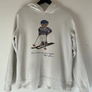 Vit hoodie Polo Bear Ralph Lauren xs/s - Vit hoodie från Polo Ralph Lauren i storlek Xs/s med ikoniskt Polo Bear-tryck på framsidan där björnen åker skidor. Tröjan har huva, långa ärmar och är tillverkad i mjuk bomullsmix. Perfekt för dig som gillar klassisk och sportig stil.