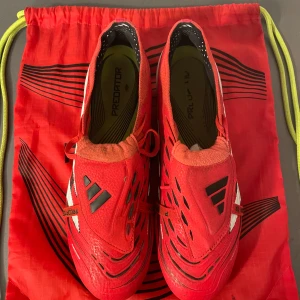 Adidas Predator röda fotbollsskor - Säljer ett par Adidas Predator fotbollsskor i rött med vita och svarta detaljer. Skorna har snörning, låg profil och mönstrad ovandel för extra grepp. Yttersulan har fasta dobbar för spel på gräs. Tillverkade i syntetmaterial och levereras med matchande röd påse.
