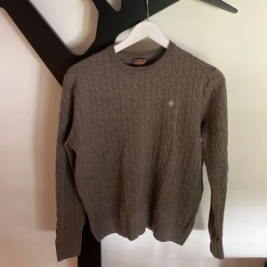 Brun kabelstickad tröja Morris L (passar S) - Säljer en brun kabelstickad tröja från Morris i storlek L, tillverkad i 100% merinoull. Tröjan har rund hals, ribbade muddar och ett litet Morris-emblem broderat på bröstet. Perfekt för dig som gillar klassisk stil med snygga detaljer. På grund av att den har krympt lite i tvätten så passar den som storlek S.