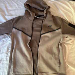 Beige Nike vindjacka med huva - Snygg beige vindjacka från Nike med svart logga på bröstet och coola svarta detaljer längs axlarna. Jackan har huva, hel dragkedja och en ficka med dragkedja på ärmen. Perfekt för dig som gillar sportig och clean stil.