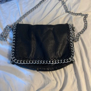 Svart axelväska med kedjedetaljer - Säljer min svarta handväska ifrån Tiamo! Den är använd och har lite defekter på vissa ställen (se bilder) annars fungerar den bra. Höjd 13cm bredd 20cm