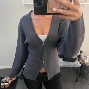 Mörkgrå cardigan från Gina Tricot - Mörkgrå cardigan från Gina Tricot med v-ringning och klädda knappar framtill. Perfekt att slänga på sig över ett linne eller t-shirt. Använd ett fåtal gånger!