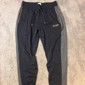 Svarta mjukisbyxor från BOSS - Snygga svarta mjukisbyxor från BOSS med grå sidopaneler och gula detaljer vid fickorna. Byxorna har dragsko i midjan och BOSS-logga på låret. Perfekta för en chill och sportig stil. Äkta och aldrig använda.