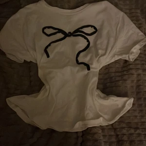 Vit t-shirt med svart rosett - Vit t-shirt i bomull med korta ärmar och en svart tryckt rosett framtill. Enkel och söt design som passar perfekt till jeans eller kjol. T-shirten har en klassisk passform och rund halsringning.