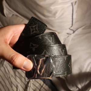 Louis Vuitton svart monogram bälte - Snyggt svart bälte från Louis Vuitton med klassiskt monogrammönster och silverfärgat LV-spänne. Bältet är gjort i skinn med en insida i ljusbrun nyans och har exklusiva detaljer som sticker ut. Perfekt accessoar för att lyfta din outfit.