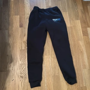 Svarta Trapstar mjukisbyxor - Svarta mjukisbyxor från Trapstar med blått och vitt tryck på ena benet där det står 'Trapstar It's a Secret'. Byxorna har resår i midjan och muddar vid bensluten för en skön och avslappnad passform. Perfekta för chill eller streetwear.